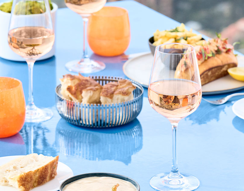 3 glasses of rose on a blue table with a selection of food, including a bread basket