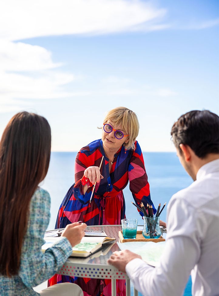 There are two people doing a watercolor class by the sea on the terrace of their suite, accompanied by a private instructor.