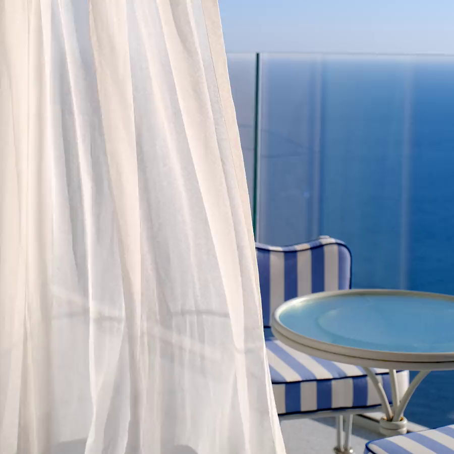 A white curtain moving next to a chair and a table with a view of the sea.