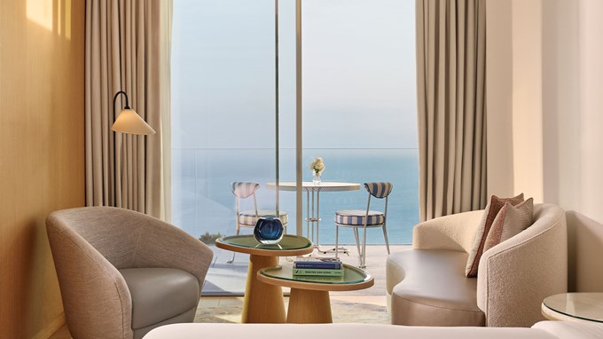 Espace salon confortable avec un mobilier moderne dans une suite, face à un balcon avec table et chaises surplombant la mer.