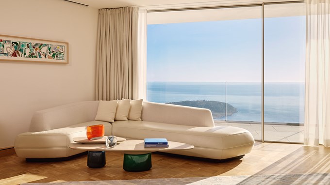 A bright living room with a curved white sofa and coffee table, featuring a floor-to-ceiling window overlooking the ocean.