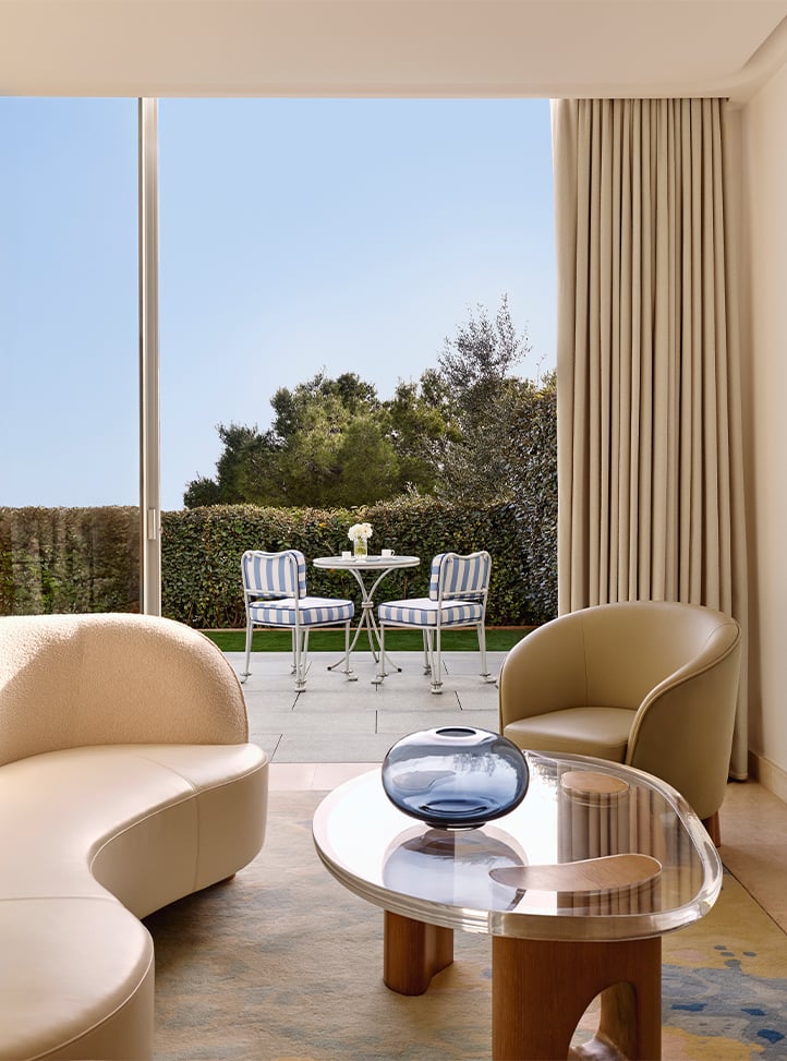 A ground-level room with modern seating and a glass coffee table, opening to a private patio with garden views and striped chairs.