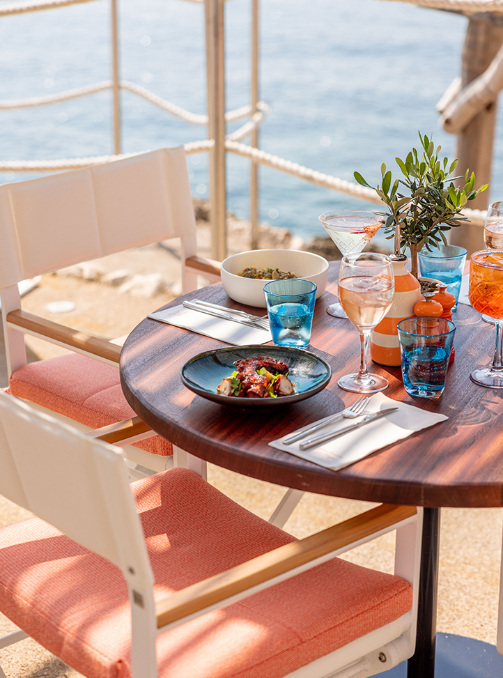 Une table à Maybourne La Plage avec des assiettes, verres et couverts et la vue sur la mer en fond.