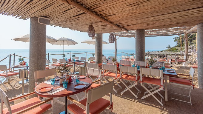 Maybourne La Plage - vue de la salle à manger avec des tables en bois rondes, des chaises blanches et corail et la vue sur la mer en fond.