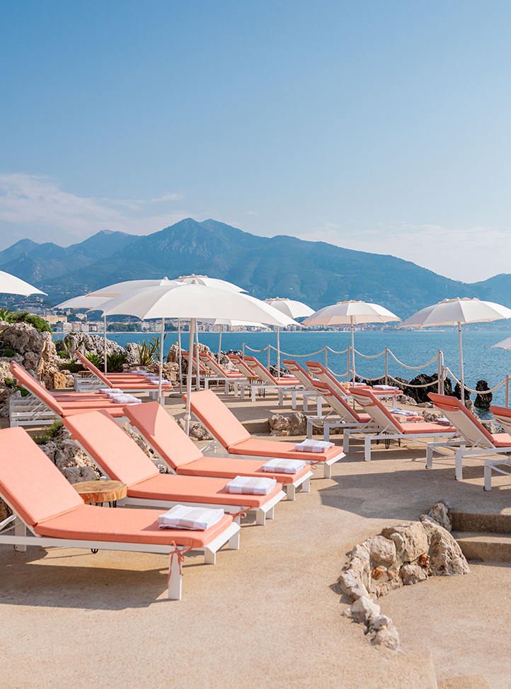 Maybourne La Plage sun loungers in a coral colour with shaders and the sea in the background.