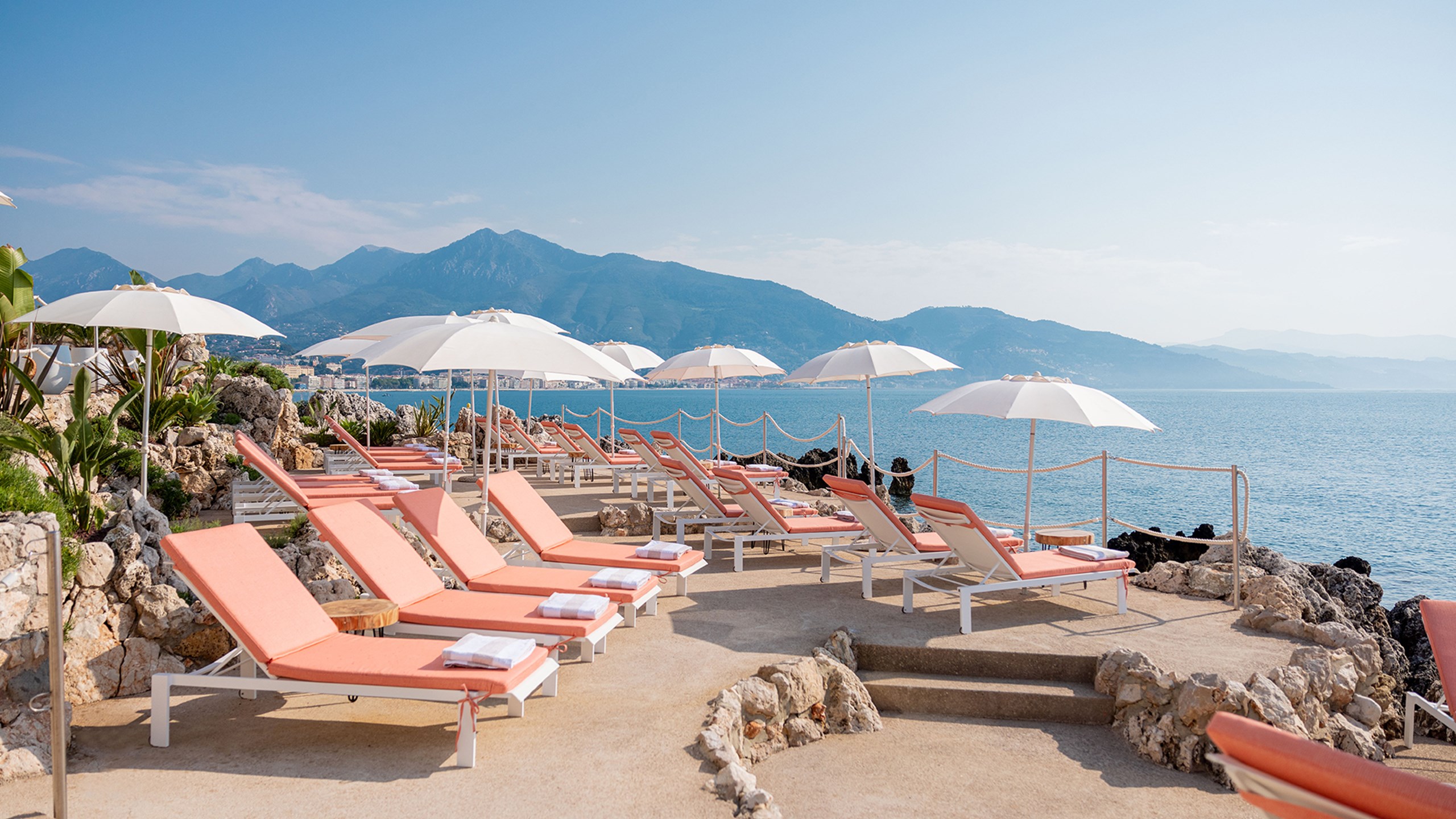 Maybourne La Plage - View of the orange - pink sun loungers with the white sun shaders, next to the sea and with the mountains in the background.
