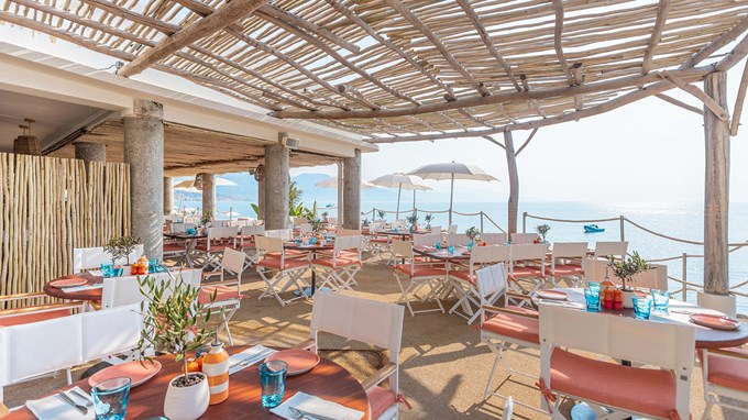 Maybourne La Plage dining area with round wooden tables, white and pink chairs and glassware on the table. There is a view on the sea in the background.