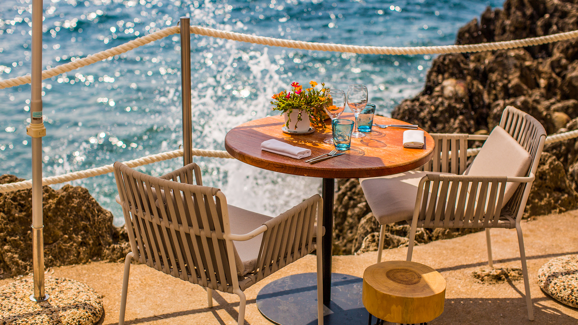 Une table avec des verres et couverts dessus, et deux chaises de part et d'autre. La mer se distingue dans l'arrière-plan.