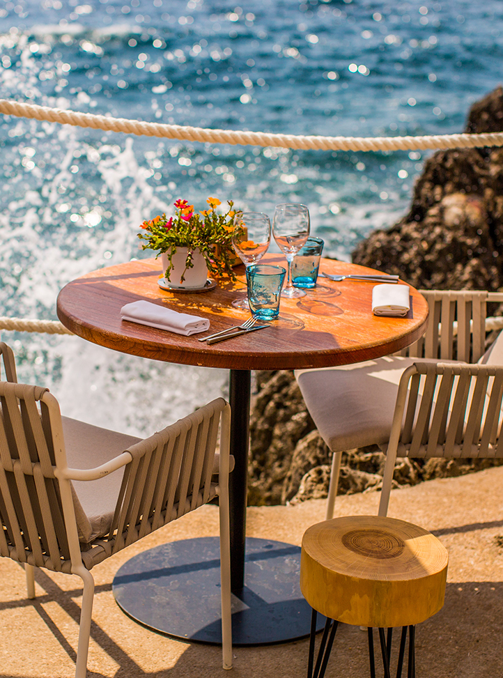 A round dining table with two chairs on each side. There are glasses and cutlery on top. The sea appears in the background.