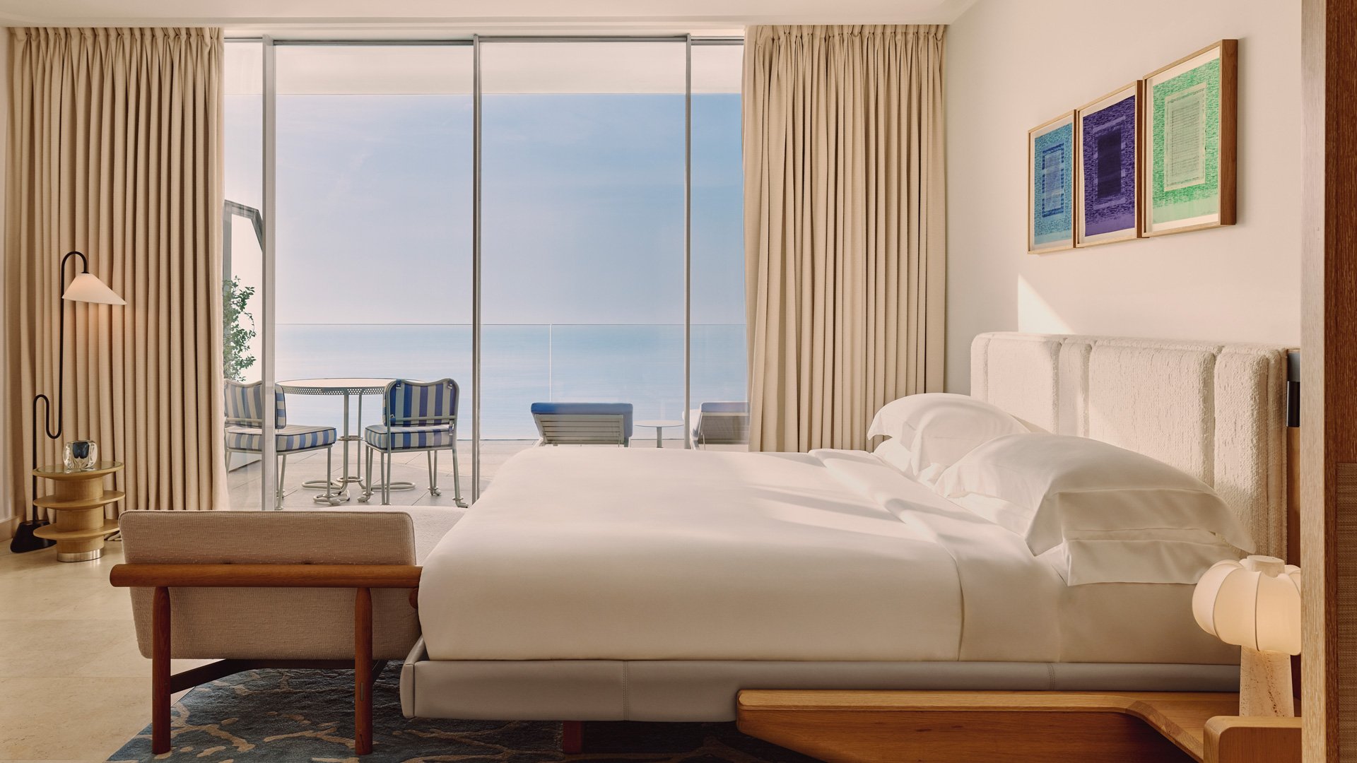 Grande chambre lumineuse avec lit double, œuvres encadrées sur le mur, rideaux beiges et accès à une terrasse avec vue sur la mer.
