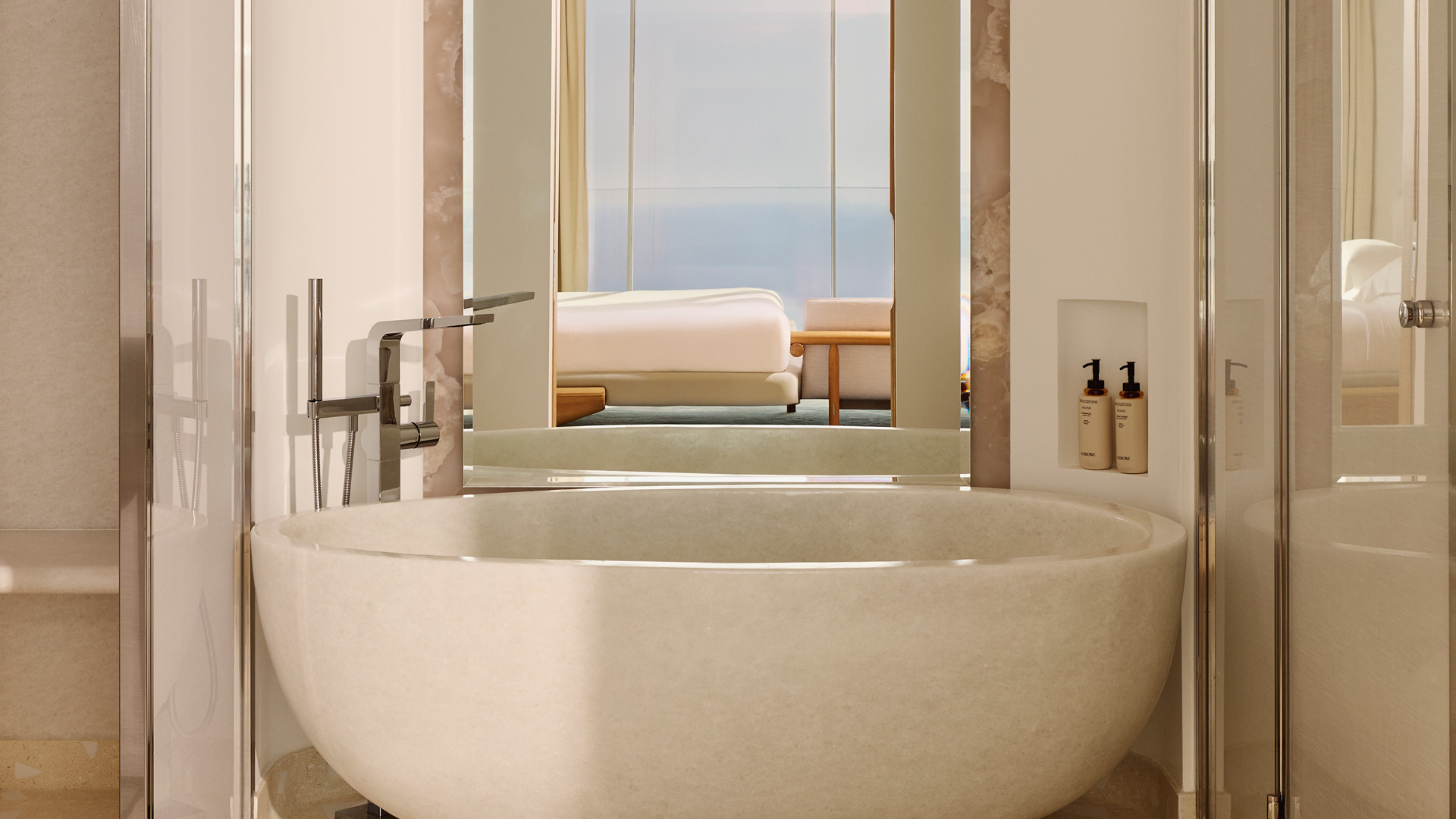 Salle de bain moderne avec vasque en pierre, robinetterie métallique, produits de toilette muraux et vue directe sur la chambre et la mer au fond.