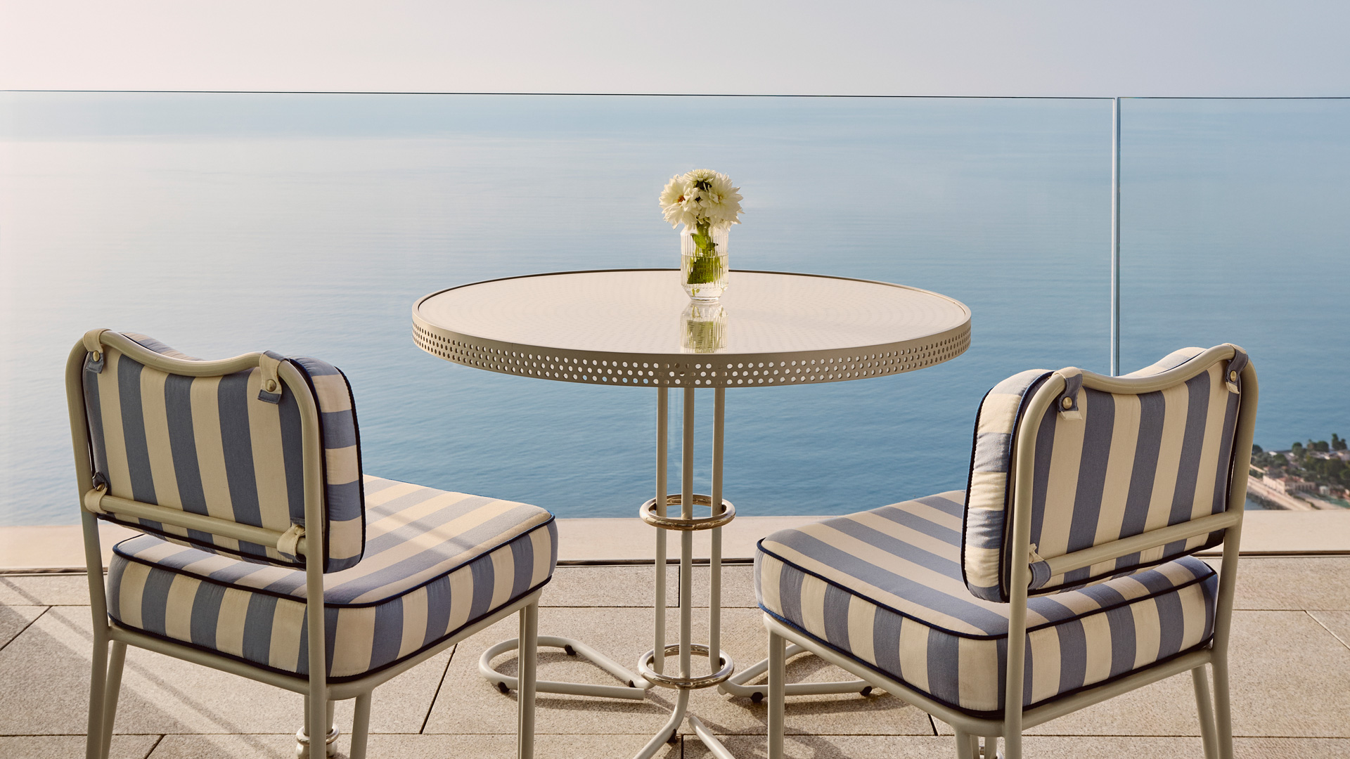 Table ronde en métal blanc avec deux chaises à rayures bleues et beiges sur une terrasse, face à une mer calme et un ciel clair.
