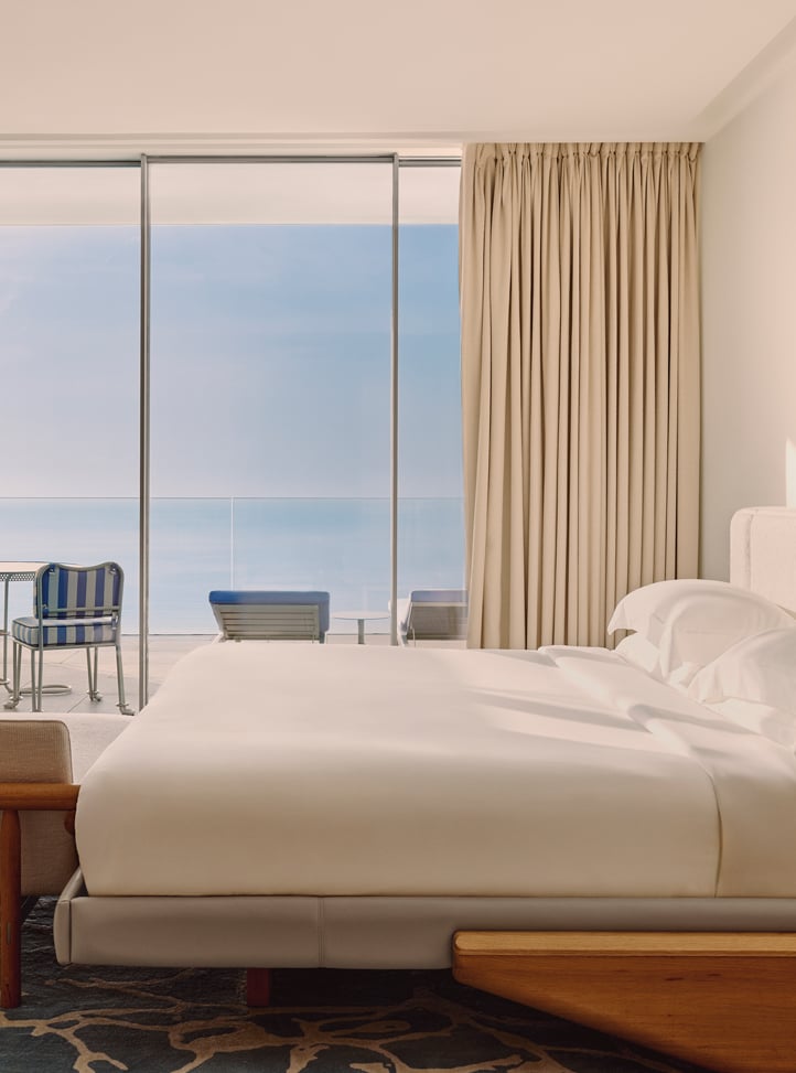 Lit double dans une chambre moderne avec vue sur une terrasse meublée et la mer à travers de grandes baies vitrées.