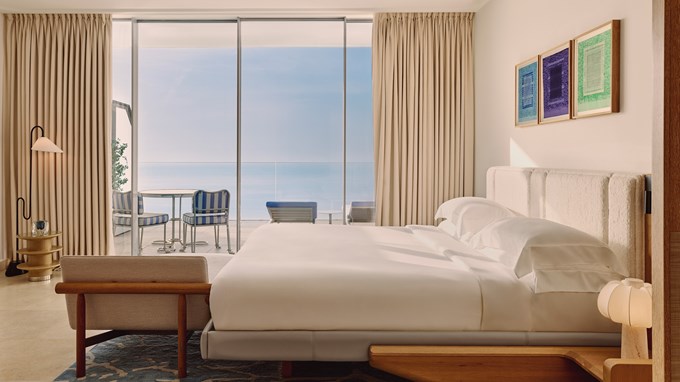 Minimalist bedroom with a low-profile bed and floor-to-ceiling glass doors leading to a sea-facing terrace.
