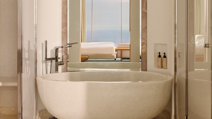 Freestanding stone bathtub in a modern bathroom with a mirror reflecting the bedroom and sea view.