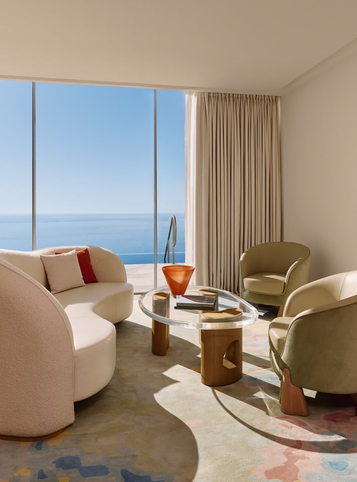 Salon lumineux avec canapé blanc incurvé, fauteuils verts, table basse en verre et grande baie vitrée donnant sur la mer.