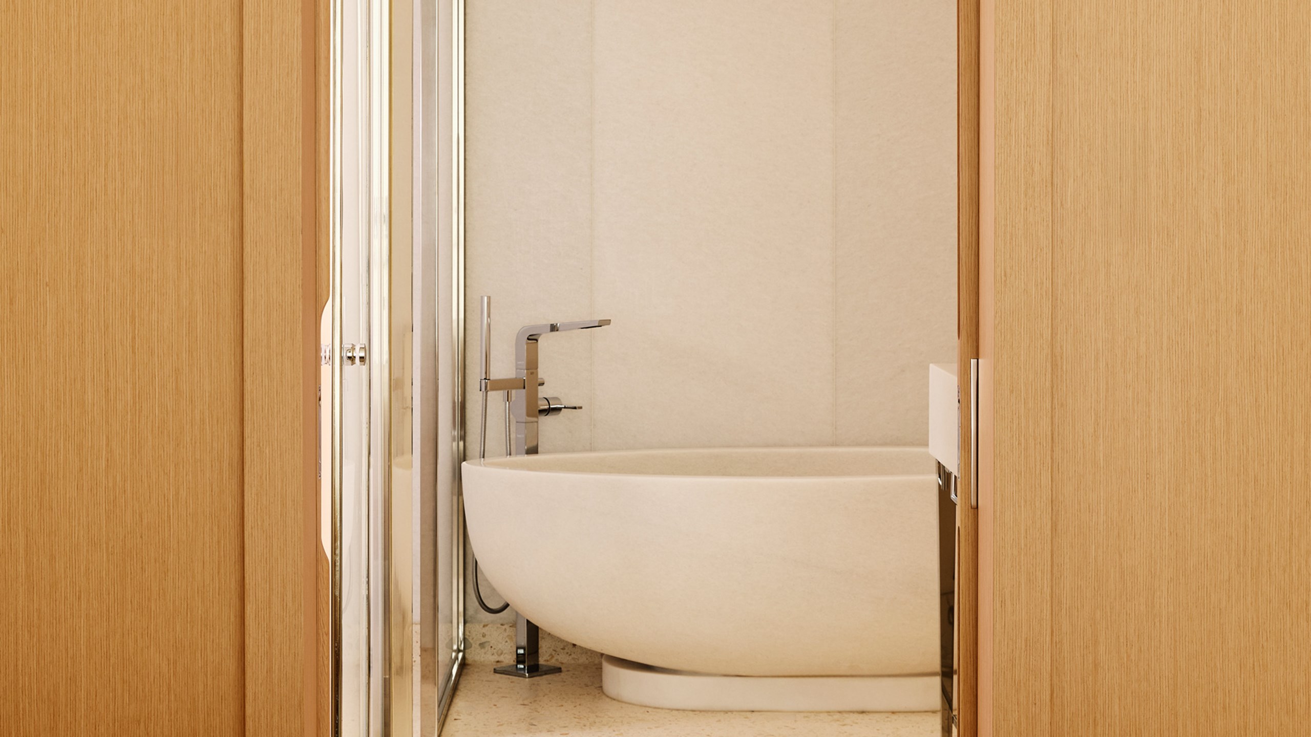Salle de bain minimaliste avec une grande baignoire en pierre beige, robinetterie design et portes en bois clair.