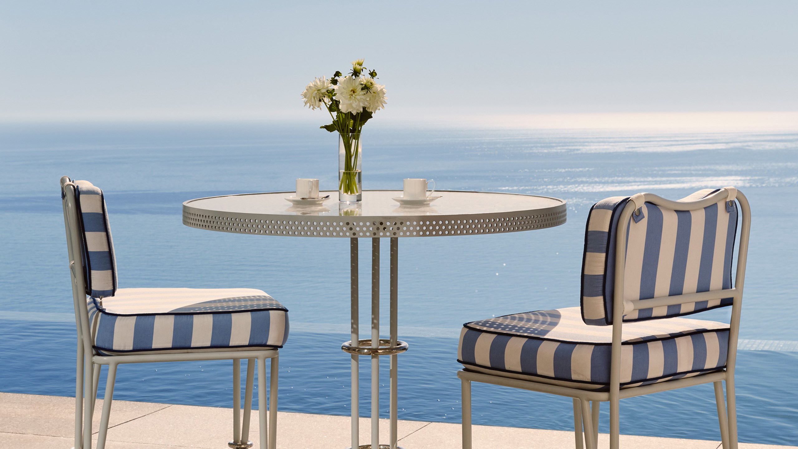 Outdoor table set for two with striped chairs and ocean backdrop.