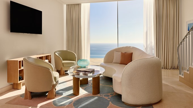 Salon moderne avec canapé incurvé, fauteuils crème, table basse en verre, télévision murale et grande baie vitrée offrant une vue sur la mer.