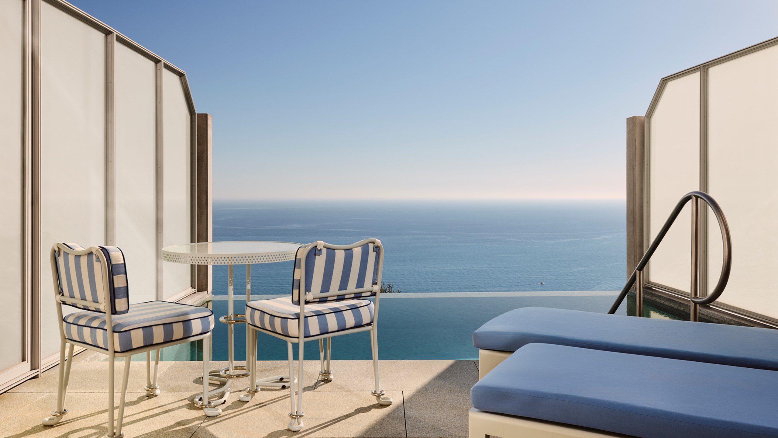 Petite terrasse privée avec deux chaises rayées bleues et blanches, une table en métal blanc et deux transats bleus face à une piscine et l’océan.