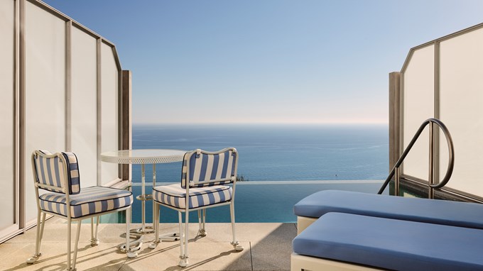 Petite terrasse privée avec deux chaises rayées bleues et blanches, une table en métal blanc et deux transats bleus face à une piscine et l’océan.