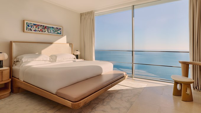 Chambre élégante avec un lit double en bois clair, décor minimaliste, œuvre colorée au mur et grande fenêtre donnant sur la mer.