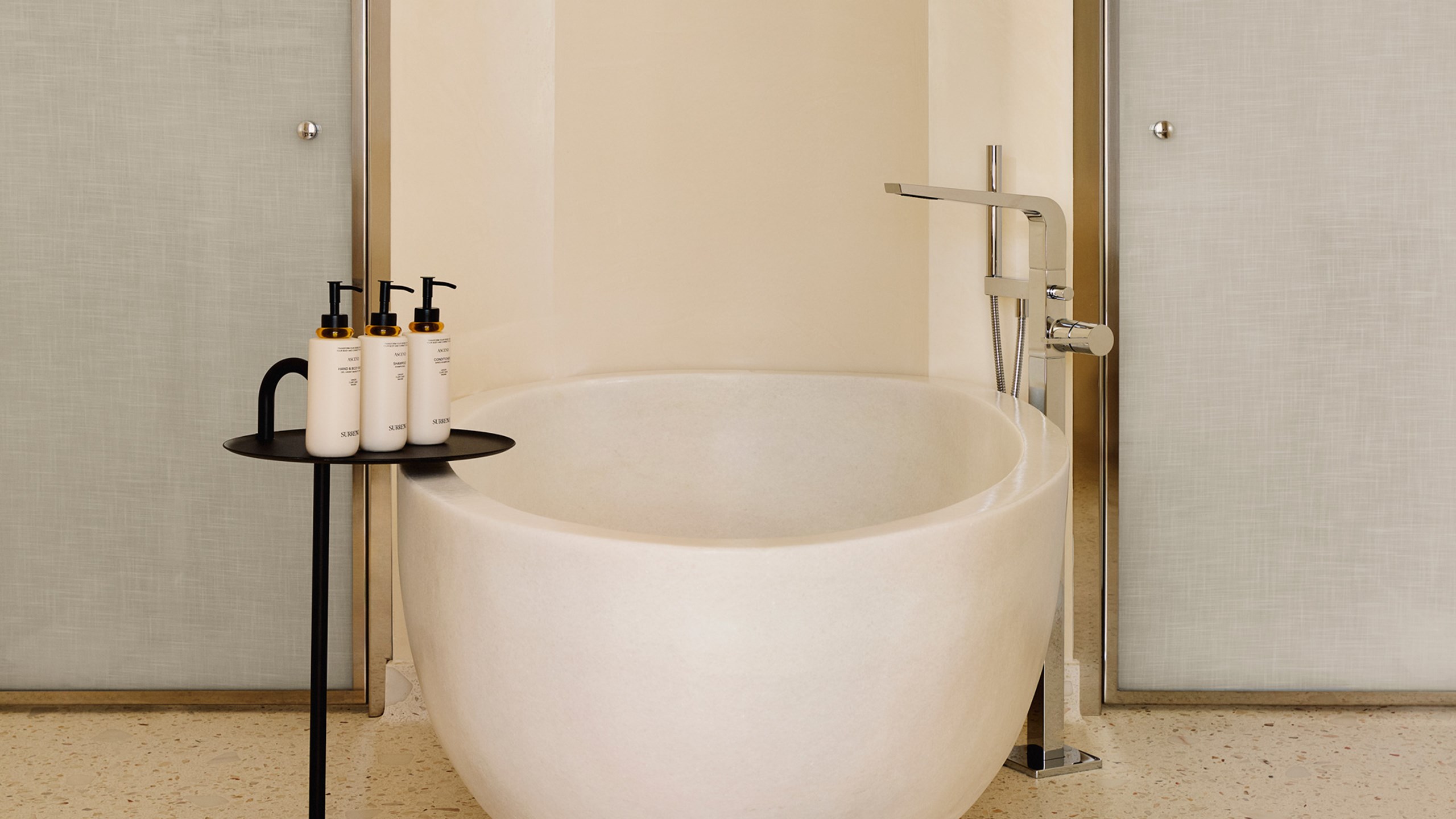 Salle de bain épurée avec baignoire en pierre blanche, robinetterie moderne et table d’appoint noire avec trois flacons de produits de toilette.