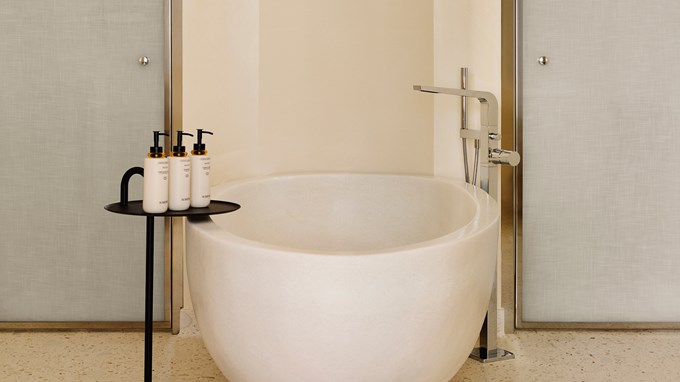 Salle de bain épurée avec baignoire en pierre blanche, robinetterie moderne et table d’appoint noire avec trois flacons de produits de toilette.