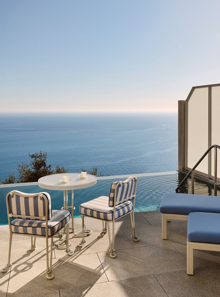 Terrasse ensoleillée avec deux chaises rayées, une petite table blanche et des transats bleus, donnant sur une piscine à débordement avec vue sur la mer.