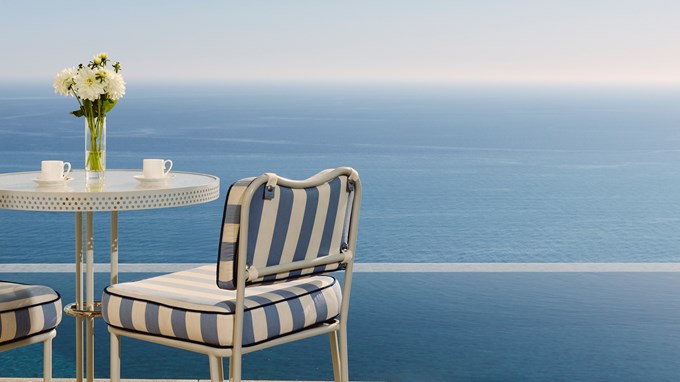 Table set for two with white cups and striped chairs facing expansive sea view.