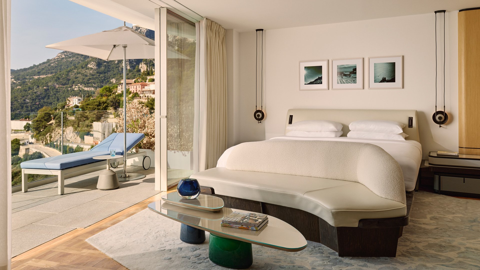 Chambre lumineuse avec lit double, mobilier contemporain et accès à une terrasse équipée d’un transat et d’un parasol, face aux collines verdoyantes.