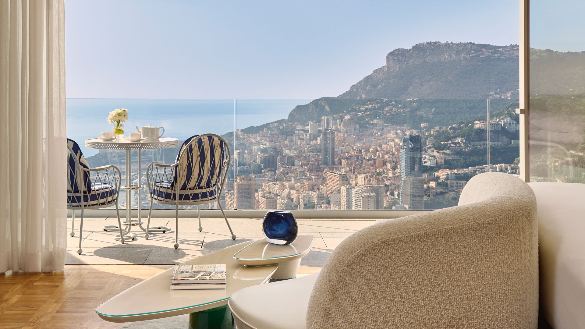 Chambre moderne avec canapé design et vue panoramique sur la ville et la mer depuis une grande baie vitrée, donnant sur une terrasse avec table et chaises.
