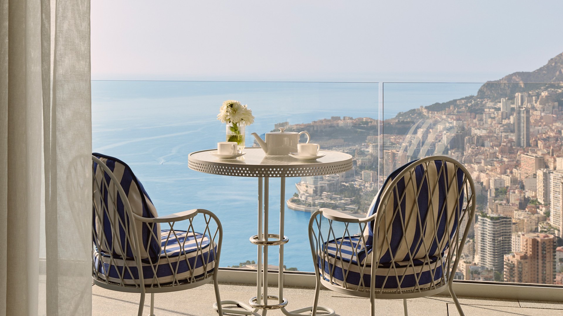 Table ronde blanche avec deux chaises rayées bleues et blanches, posée sur une terrasse surplombant Monaco et la mer Méditerranée.