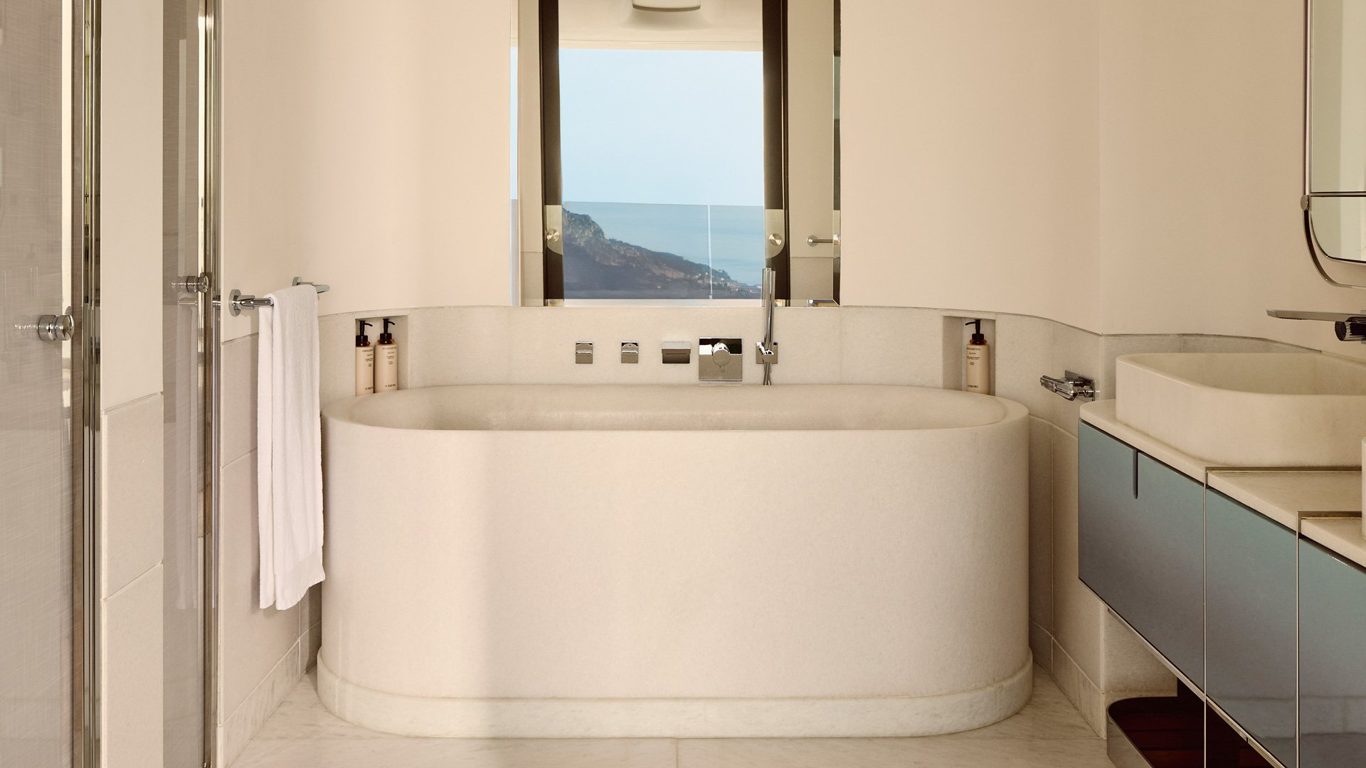Salle de bain élégante avec une grande baignoire en pierre claire, robinetterie moderne, double vasque et vue sur la mer à travers la fenêtre.