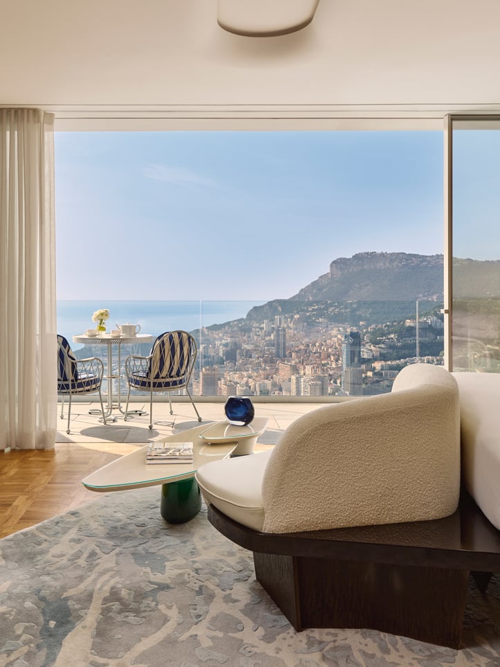Chambre moderne avec canapé design et vue panoramique sur la ville et la mer depuis une grande baie vitrée, donnant sur une terrasse avec table et chaises.