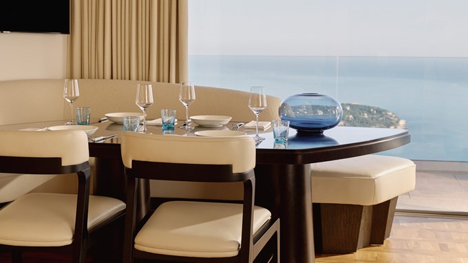 Salle à manger contemporaine avec table en bois sombre, vaisselle élégante, banquette courbée et vue panoramique sur la mer et le littoral.