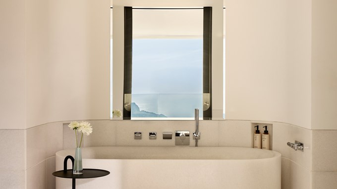 Salle de bain élégante avec baignoire en pierre blanche, robinetterie chromée, table d’appoint noire avec fleurs et vue sur la mer à travers une grande fenêtre.