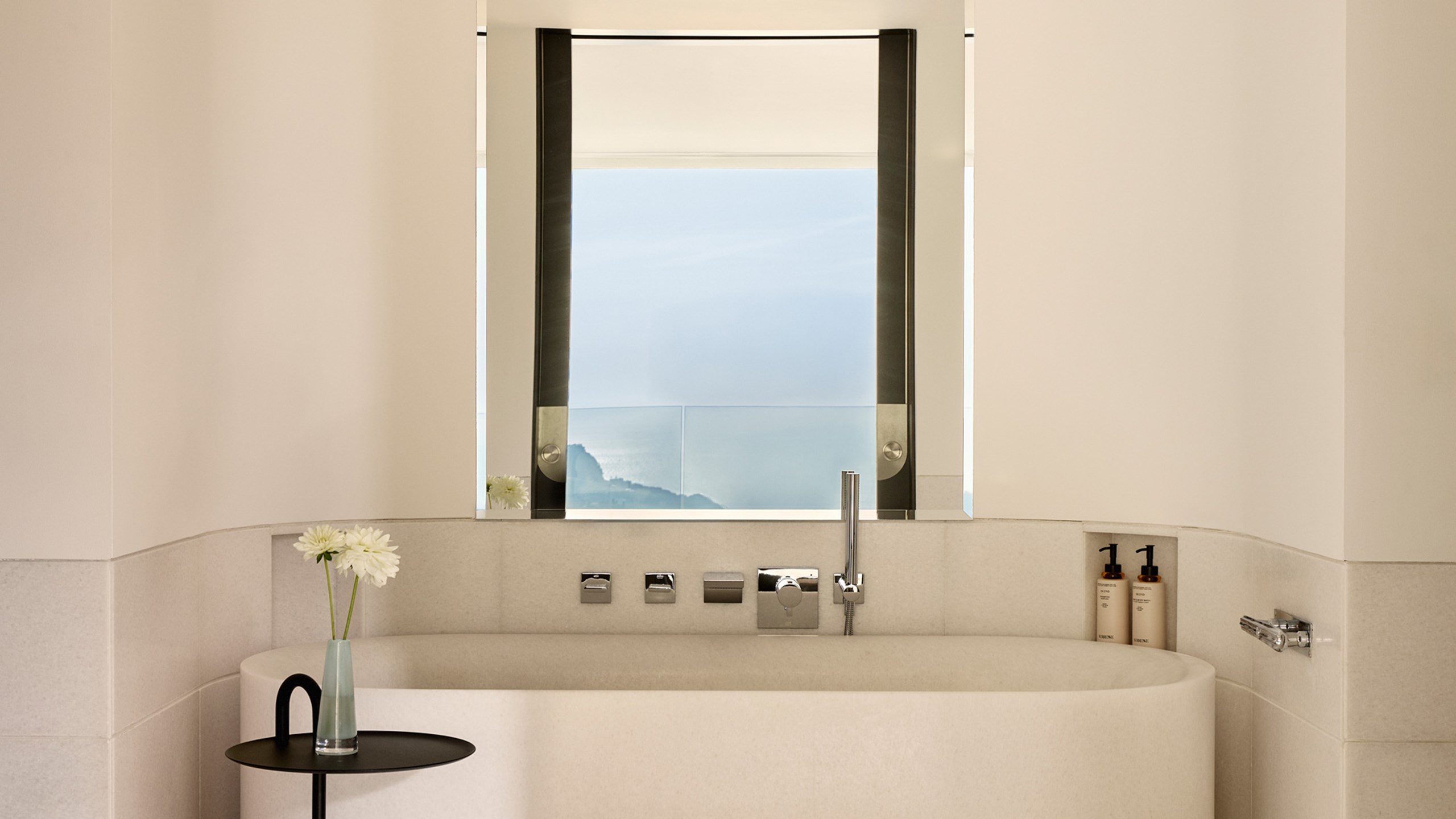 Minimalist bathroom with a freestanding tub and ocean view through tall window.
