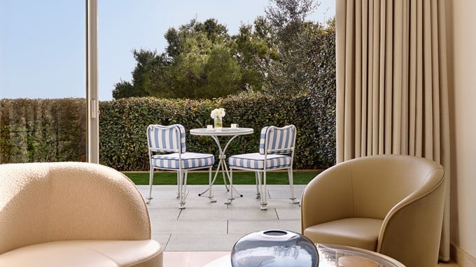 Vue d’un jardin privé avec une terrasse aménagée, deux chaises rayées bleues et blanches autour d’une table ronde, prise depuis l’intérieur d’un salon élégant.