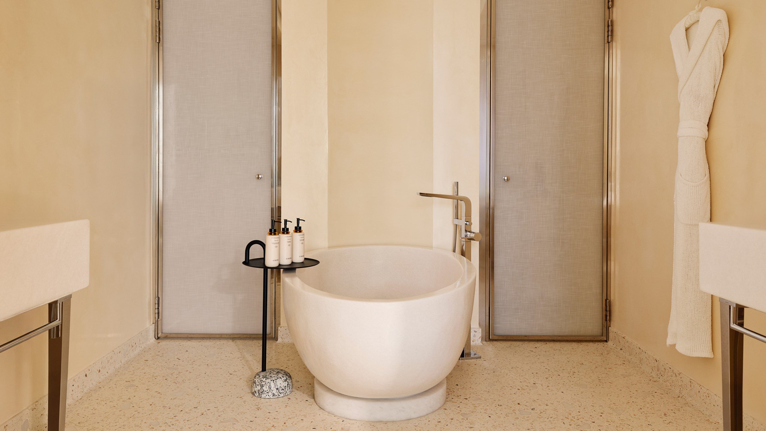 Salle de bain luxueuse avec une grande baignoire ronde en pierre, des produits de toilette alignés, des portes vitrées satinées, et un peignoir accroché au mur.