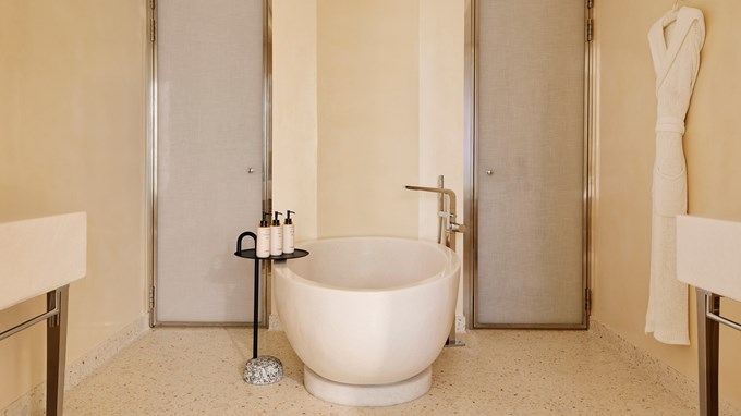 Salle de bain luxueuse avec une grande baignoire ronde en pierre, des produits de toilette alignés, des portes vitrées satinées, et un peignoir accroché au mur.