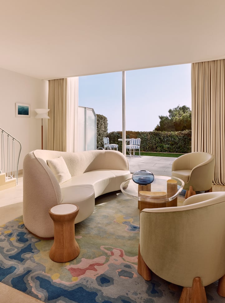 Salon lumineux et moderne avec un canapé crème incurvé, deux fauteuils beiges, une table basse en verre, et une vue sur un jardin verdoyant à travers une baie vitrée.