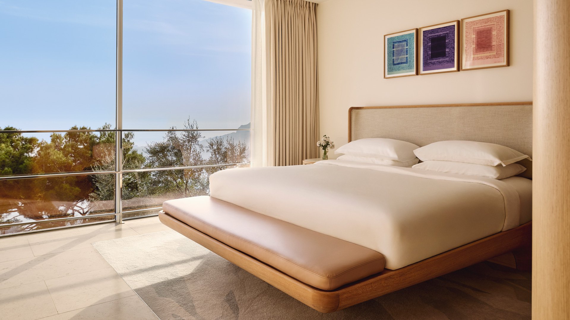Minimalist bedroom with floor-to-ceiling windows overlooking trees and a distant sea view.