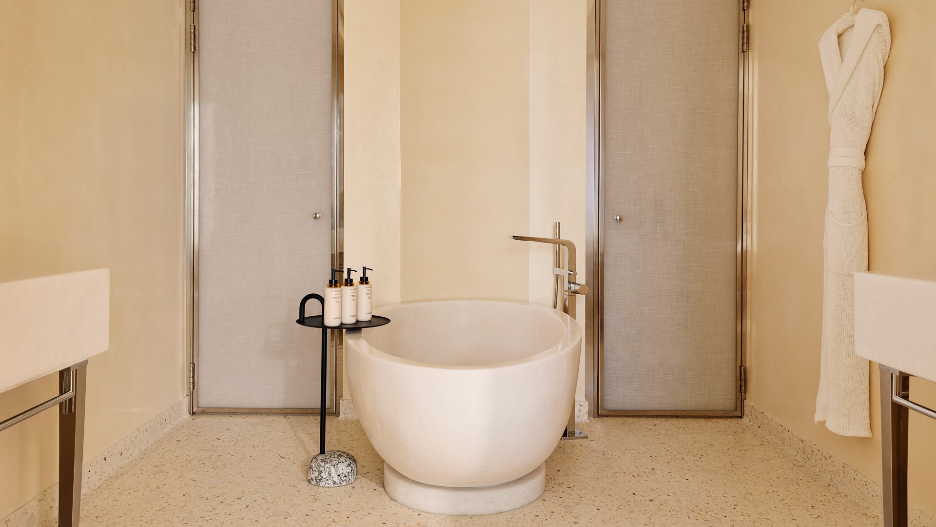 Freestanding oval bathtub in a neutral-toned bathroom, flanked by two doors and a robe on the wall.