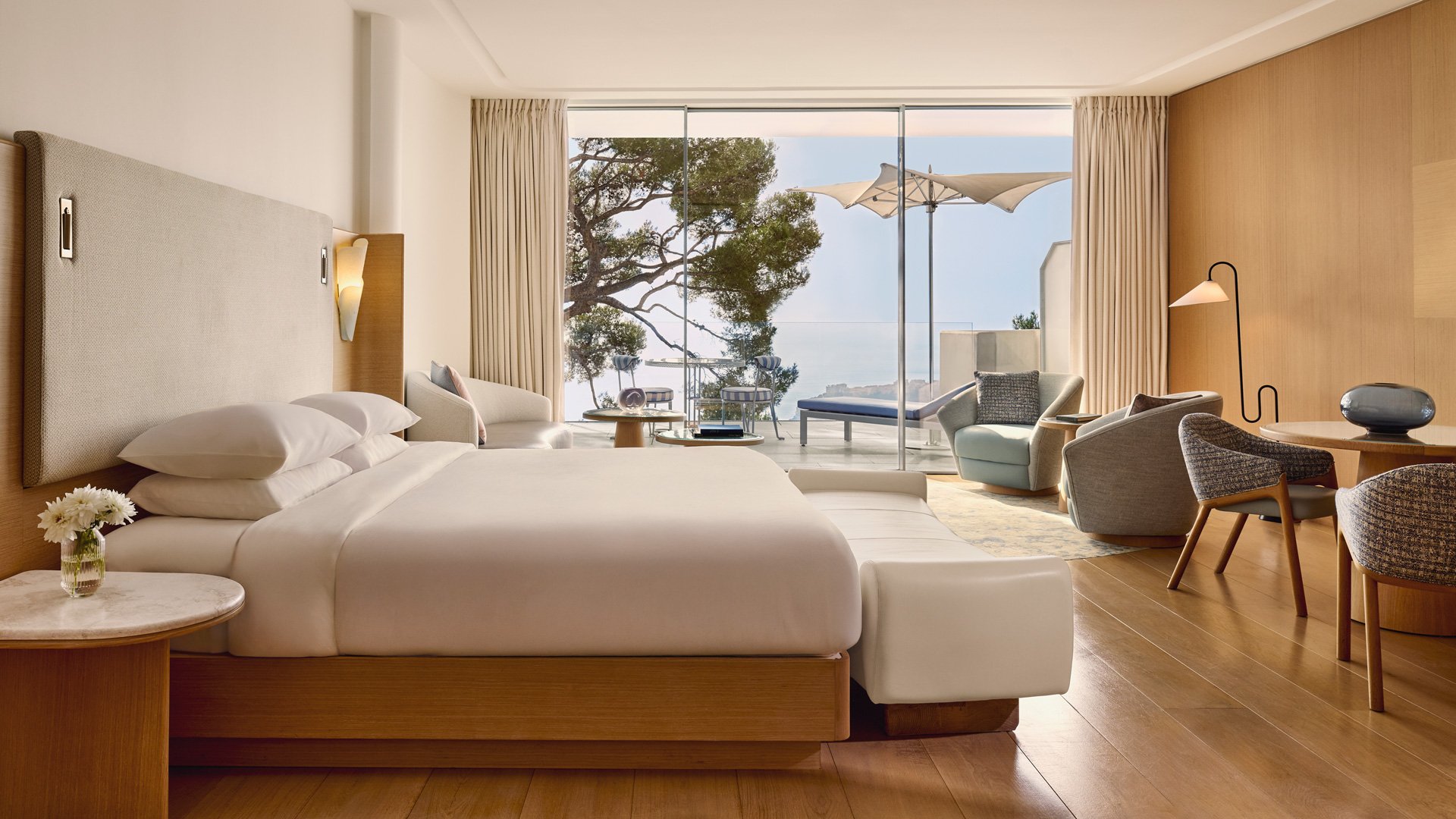 Vue d’ensemble de la suite avec lit double, coin salon, table à manger, et grande baie vitrée s’ouvrant sur une terrasse avec vue sur la mer et les pins.