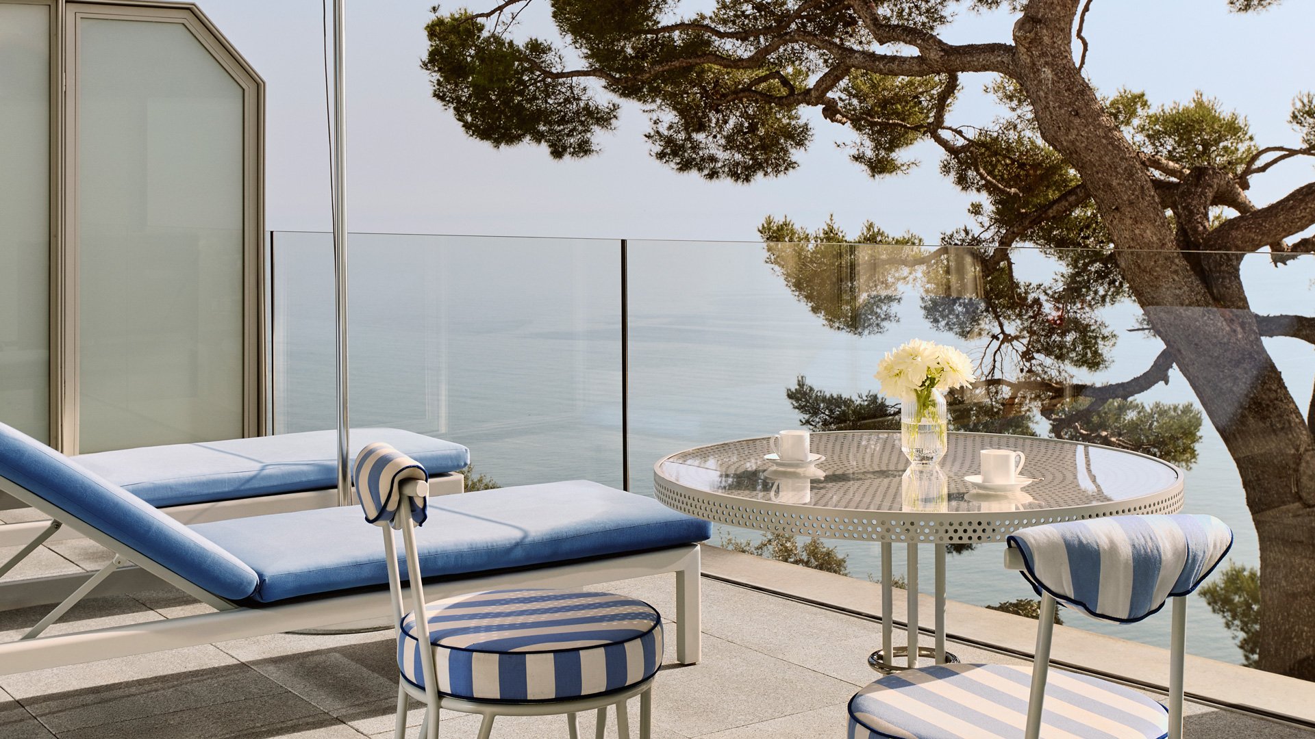 Terrasse privative avec chaises longues et table ronde, offrant une vue sur la mer et entourée d’arbres méditerranéens.
