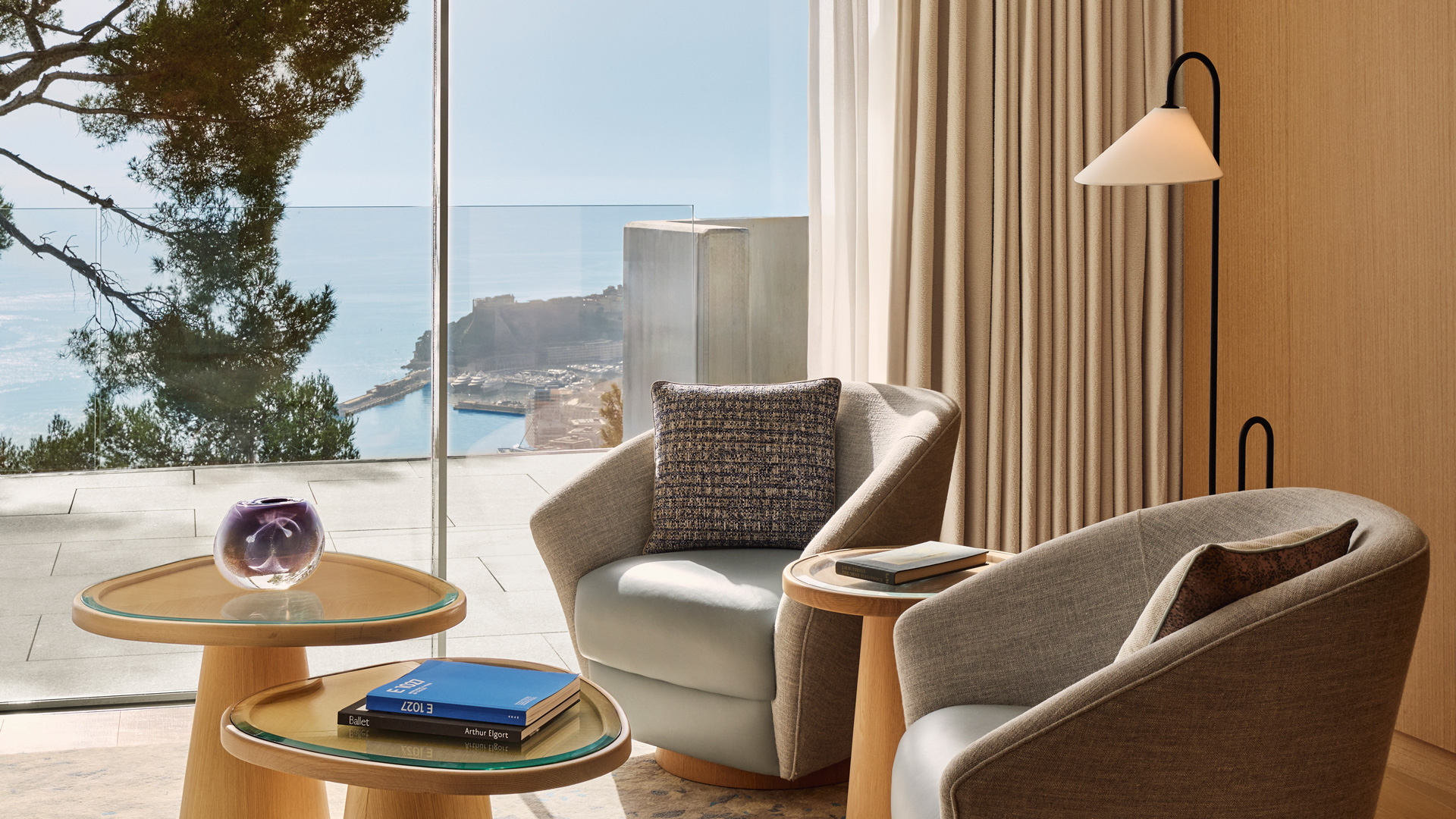 Espace salon lumineux avec deux fauteuils gris modernes, tables basses en bois clair et vue panoramique sur le port depuis une baie vitrée.