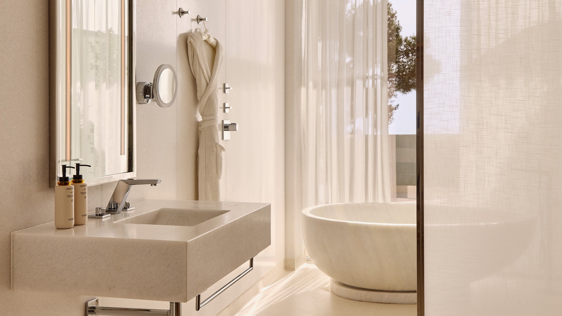 Salle de bains élégante avec vasque en pierre, miroir rétroéclairé, peignoir suspendu et baignoire ovale près d’un rideau transparent laissant filtrer la lumière naturelle.