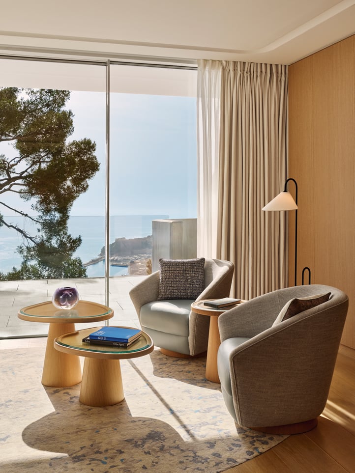 Autre vue du salon avec fauteuils gris et tables basses, face à une baie vitrée donnant sur la mer et les collines monégasques.