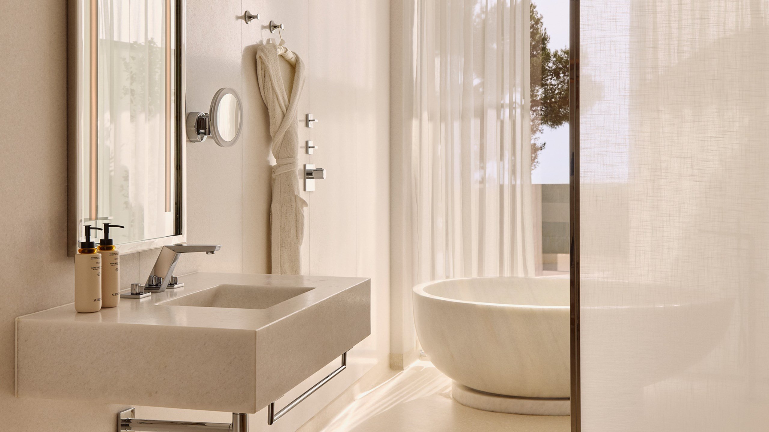 Light-filled bathroom with stone basin and freestanding tub, framed by sheer curtains and outdoor views.